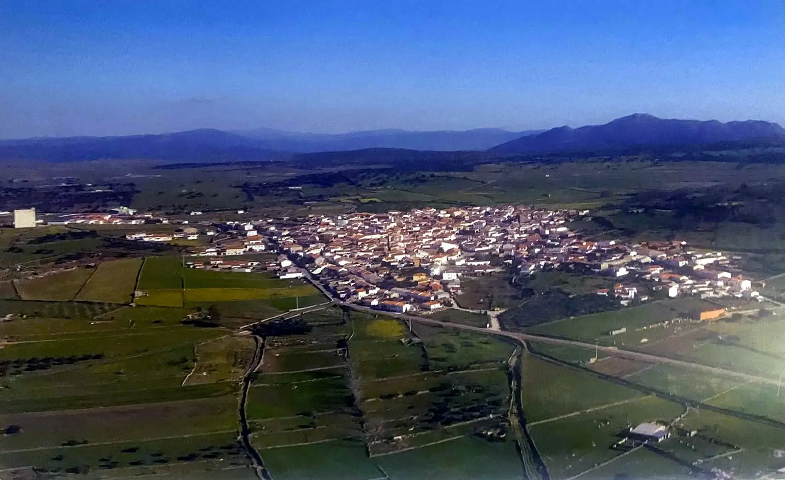 Fotografía aérea de Ibahernando: casas blancas con tejados rojos entre prados verdes, con montañas al fondo.