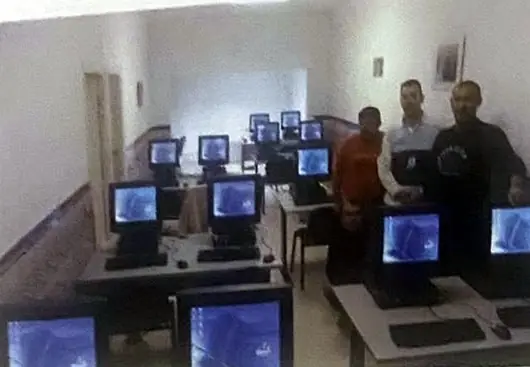 Aula con filas de ordenadores encendidos y cuatro personas al fondo.