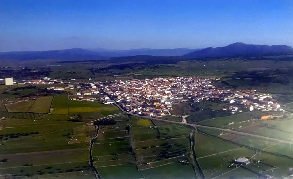 Fotografía aérea de Ibahernando: casas blancas con tejados rojos rodeadas de prados verdes y muros de piedra, montañas al fondo bajo un cielo azul.