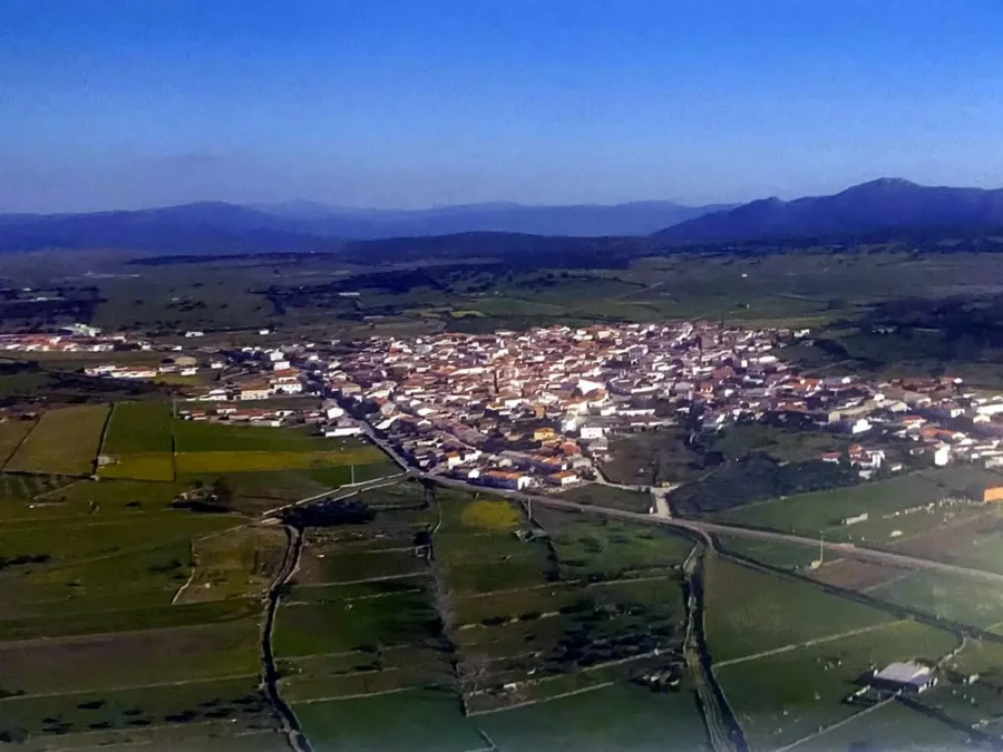 Fotografía aérea de Ibahernando: casas blancas con tejados rojos rodeadas de prados verdes y muros de piedra, montañas al fondo bajo un cielo azul.