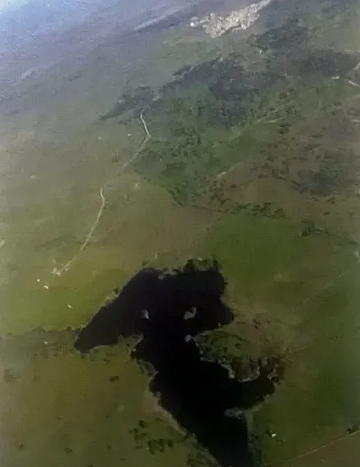 Fotografía aérea vertical del pantano, una mancha oscura de agua entre dehesa verde, con el pueblo a lo lejos.