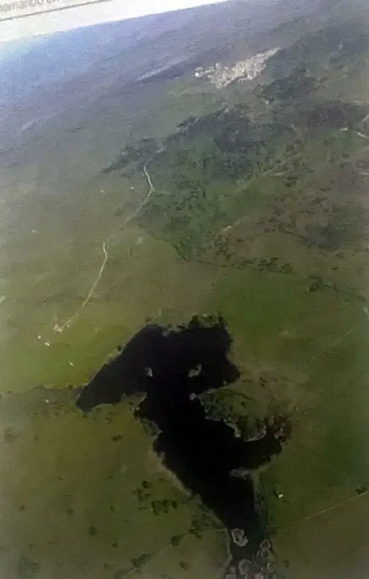 Fotografía aérea vertical del pantano, una mancha oscura de agua entre dehesa verde, con el pueblo a lo lejos.