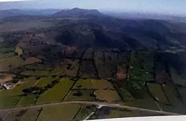 Panorámica aérea de cercados verdes que suben hacia la sierra, con la luz baja de la tarde.