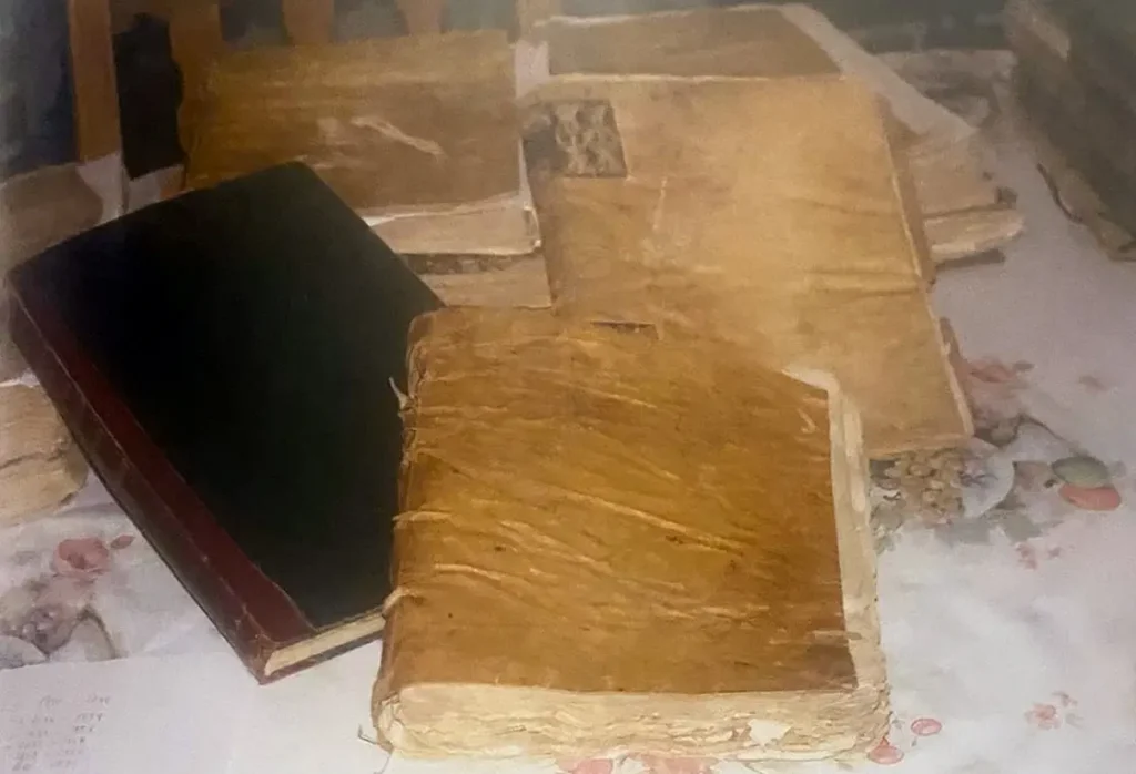 Varios libros antiguos encuadernados en pergamino amarillento, uno abierto, sobre un mantel de flores.