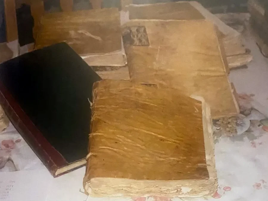 Varios libros antiguos encuadernados en pergamino amarillento, uno abierto, sobre un mantel de flores.