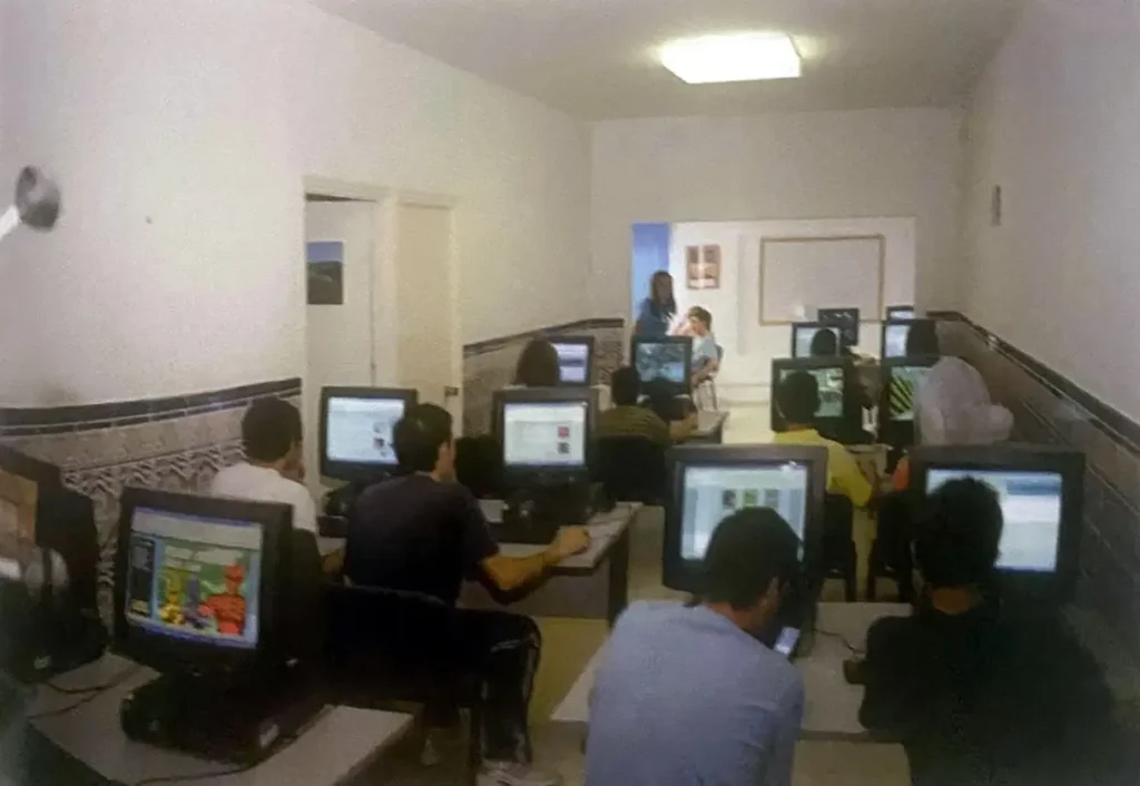 Aula de paredes blancas y zócalo de azulejo con una docena de personas trabajando ante ordenadores.