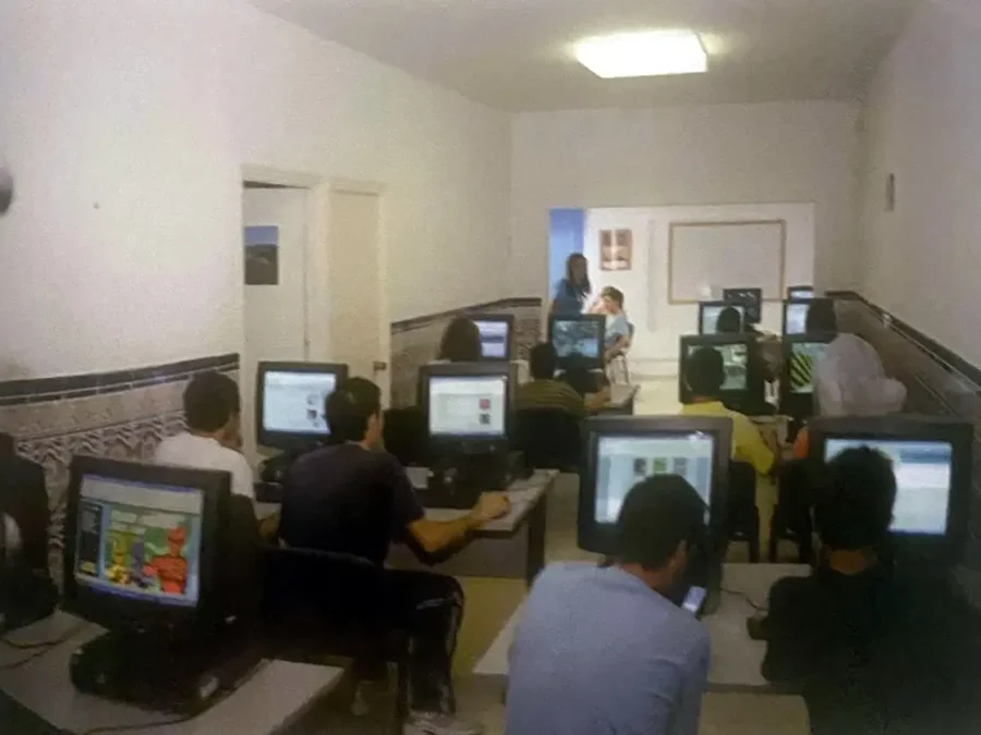 Aula de paredes blancas y zócalo de azulejo con una docena de personas trabajando ante ordenadores.