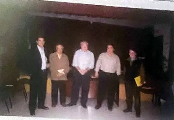 Seis hombres de pie en fila sobre un escenario con telón oscuro.