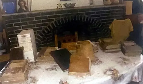 Mesa con libros parroquiales antiguos apilados y cajas de archivo, ante una chimenea de ladrillo.