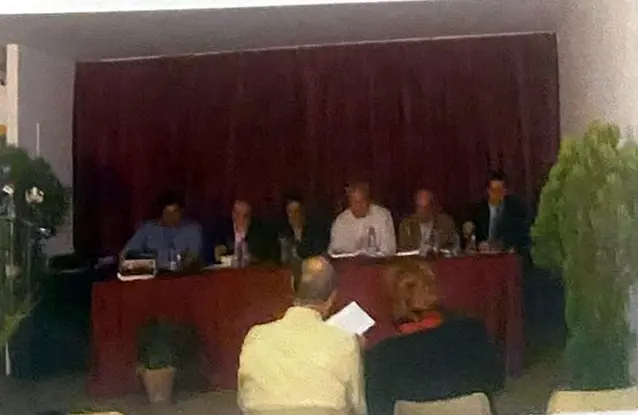 Mesa presidencial con mantel granate y varias personas sentadas, con público en primer plano.